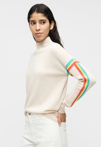 Μπεζ πουλόβερ με ψηλό λαιμό, με λωρίδα σε χρώματα του ουράνιου τόξου στο αριστερό μανίκι. Μαλακή υφή και άνετη εφαρμογή. Συνδυασμένο με λευκό παντελόνι.