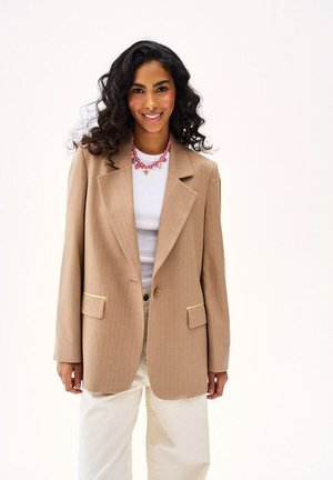 Kobieta z kręconymi czarnymi włosami, ubraną w beżową marynarkę na białej koszuli i jasnych spodniach, uśmiechająca się na tle jasnego, jednolitego tła.