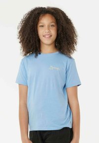 Niño con cabello rizado que lleva una camiseta de Rip Curl de color azul claro y pantalones negros, sonriendo frente a un fondo blanco liso.