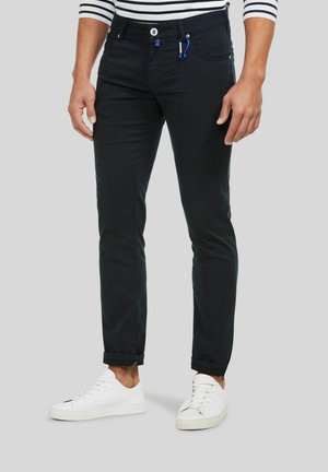 Man draagt strakke zwarte broek met opgerolde pijpen, een gestreept overhemd, witte sneakers en een blauwe karabiner vastgeklikt aan de voorzak.