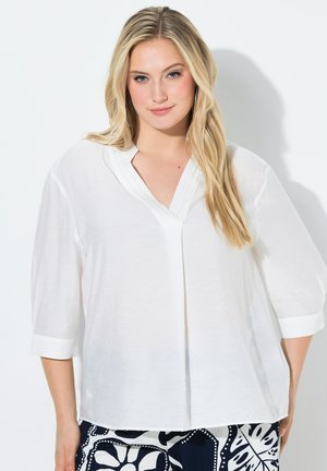 Vaalea nainen pukeutuneena löysään valkoiseen puseroon, jossa on kolme neljäsosahihnaa, ja kuvioituun mustavalkoiseen hameeseen valkoisen taustan edessä.