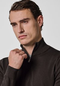 Suéter marrón claro con cremallera, cuello alto, puños acanalados y textura suave, usado sobre una camisa gris con detalles sutiles.