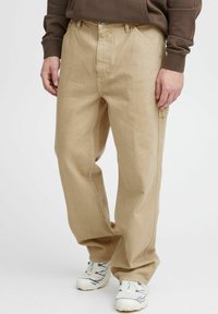 Beige bomuldsbukser med lige snit, der har dybe lommer og synlige sømme. Sammen med hvide og grå sneakers.
