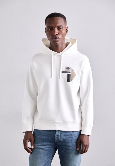 Weißer Hoodie mit Kängurutasche, Kapuze mit Kordelzug und strukturierten Patches in kontrastierenden Farben. Kombiniert mit blauen Jeans.