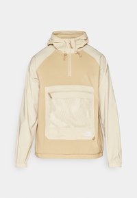 Anorak beige con diseño de media cremallera, bolsillo de malla, puños elásticos y texturas de tejido en contraste. Logo exhibido en la parte inferior derecha.