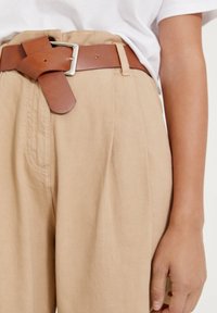 Pantalons beiges taille haute avec plis, associés à une ceinture en cuir marron avec une boucle rectangulaire en argent. Texture de tissu lisse.