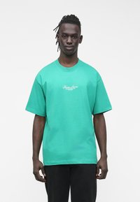 Zelené bavlněné tričko s kulat�ým výstřihem, krátkými rukávy a volným střihem. Na hrudi je vytištěno bílé logo "Guess Jeans."