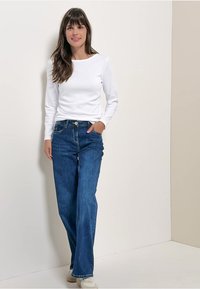 Weißes Langarmhemd, blaue gerade Jeans und weiße Sneaker. Glatter Stoff, lockerer Schnitt, mit Fronttaschen und einem einfachen Design.