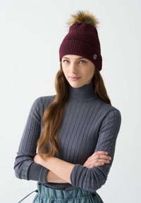 Burgundijas adīts beanie ar mākslīgo kažoku pom-pomu, ar ribotu tekstūru un logotipa akcentu. Modelis ģērbies pieguļošā pelēkā ribotā augstākajā apkaklē.