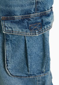 Denim cargo-housut vaaleansinisellä pesulla, joissa on suuri etutasku, näkyvät saumat ja rento leikkaus toiminnallisuutta varten.