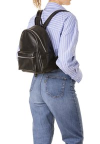 Zaino in pelle nera con due cerniere e una tasca frontale, indossato sulla schiena. Camicia a righe blu e jeans di denim visibili. Spallacci regolabili.