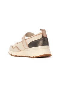 Zapatilla con parte superior suave en color beige, acento en el talón gris oscuro y correa ajustable. Presenta una suela blanca gruesa con un relieve marrón.