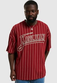 T-shirt oversize a righe rosse con un collo a costine. Presenta il logo "KARL KANI" in crema e il testo "EST.1989" sopra di esso. Materiale in cotone.
