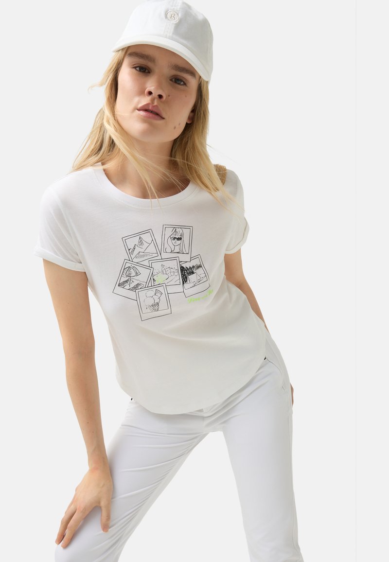 White cotton t-shirt with rolled sleeves, featuring a graphic of black-and-white photo frames and green text. Worn with white pants and a cap.