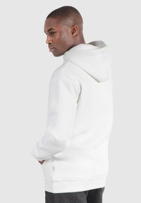 Haut à capuche blanc avec une texture douce, doté d'une capuche à cordon de serrage et d'une poche kangourou, associé à un pantalon de jogging gris. Modèle montré de dos.