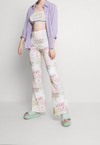 Multikleurige flared broeken met bloem- en geometrische patronen, gecombineerd met een bijpassende crop top en een lichtpaarse knoopblouse. Groene platform sandalen.