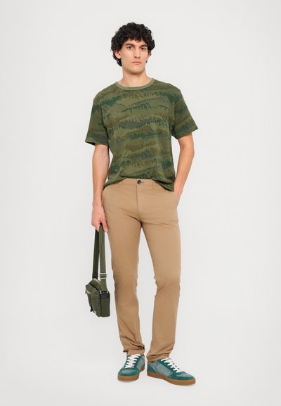 T-shirt verde camouflage, pantaloni slim-fit beige, sneakers verdi e blu, che porta una piccola borsa verde. Design semplice e look casual.