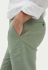 Pantalon en coton vert clair avec poches latérales et poche arrière passepoilée. Texture lisse, silhouette ajustée. Porté avec une chemise en lin blanche.
