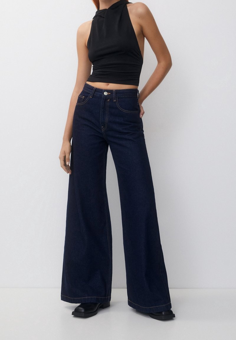 Jean en denim foncé à jambes larges avec des coutures contrastées, taille ajustée et taille haute, associé à un top halter noir. Tissu texturé.