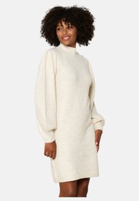 Abito in maglia color crema con collo alto e maniche lunghe a sbuffo. Tessuto testurizzato con polsini e orlo a costine. Dettagli di pattern sottili.