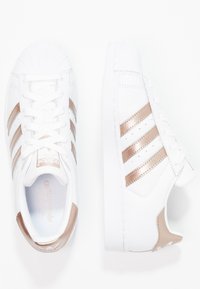 Sneakers bianchi Adidas con righe metalliche oro rosa, design con punta a conchiglia e lacci, mostrati da viste dall'alto e di lato su uno sfondo bianco.