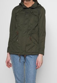 Kurtka w kolorze oliwkowej zieleni z kapturem, zamknięciem na zamek i sznurkami do regulacji. Posiada dwie przednie kieszenie oraz miękką teksturę, wykonana z trwałego materiału.