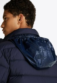 Marineblauwe gewatteerde jas met een glanzende, gestructureerde capuchon. Bevat een gestikt ontwerp en elastische gathering op de rand van de capuchon.