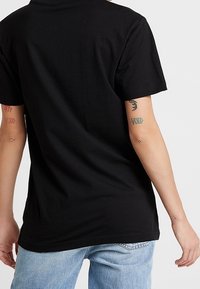 Czarny bawełniany t-shirt z okrągłym dekoltem i krótkimi rękawami, noszony z jasnoniebieskimi dżinsami, ukazujący subtelne tatuaże na ramionach.