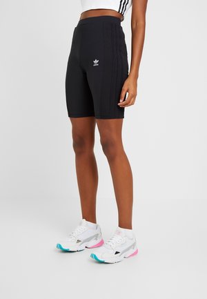 Osoba nosząca czarne rowerowe spodenki Adidas z logo na udzie oraz białe trampki z różowymi i niebieskimi akcentami na jednolitym tle.