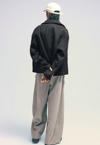 Black oversized jacket and loose gray trousers. The model wears a white cap with a logo, hands clasped behind the back. Smooth, warm textures.