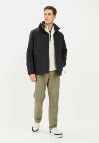 Donkerblauwe waterdichte jas met een capuchon, met twee voorzakken, twee ritsen en een gestructureerde afwerking, gedragen met khaki broek en sneakers.