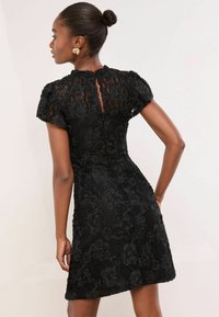 Rochie neagră din dantelă, cu texturi florale, mâneci scurte tip capă, croială ajustată și închidere cu nasturi la gât, cu tiv ondulat.