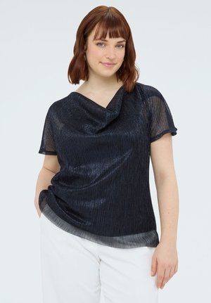 Frau mit kurzem kastanienbraunem Haar, die einen durchsichtigen schwarzen Kurzarm-Oberteil und eine weiße Hose trägt, steht mit einer Hand in der Tasche vor einem hellen Hintergrund.