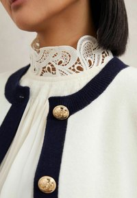 Bela jopica z mornarsko modrim robom, ki ima visok čipkast ovratnik in zlate gumbe z vgraviranimi vzorci. Mehka tekstura in klasična zasnova.