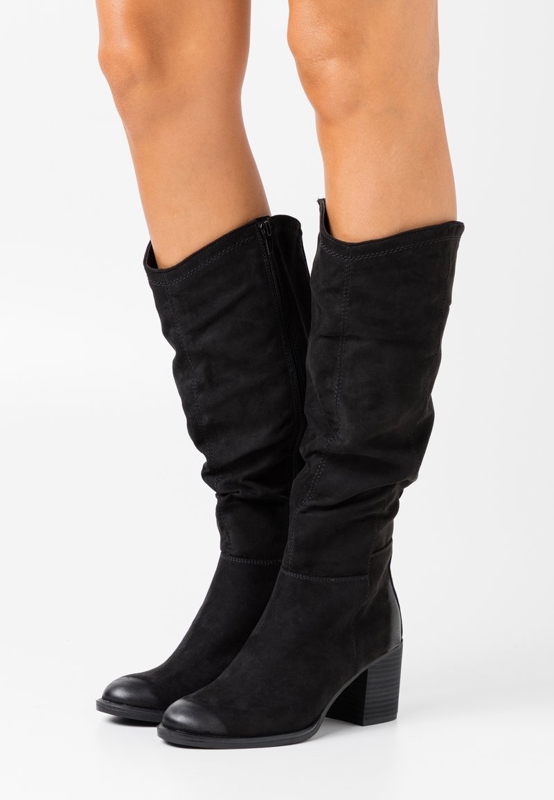 Bottes hautes en daim noir jusqu'aux genoux avec talons blocs, portées sur jambes nues, avec des fermetures éclair latérales et une texture légèrement froissée de la tige.