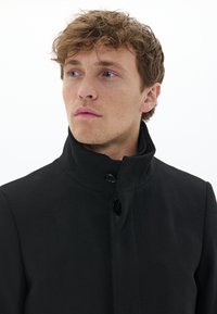 Jeune homme aux cheveux bouclés châtain clair portant un manteau noir à col montant, regardant pensivement vers la gauche sur fond blanc.