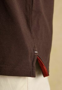 Brūns polo krekls ar teksturētu apdari, ar sānu šķēlumu un sarkanu akcentu apakšmalā. Šuvju vīles ir redzamas gar malām.