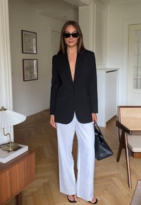 Juodas švarkas su giliu V formos iškirpimu, derinamas su baltais plačiais kelnais, kurių apačioje yra aklų detalių, ir juodais sandalais. Juodas rankinukas ir saulės akiniai.