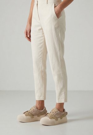 Person iført lyse beige slim-fit bukser og beige sneakers med tykke såler, stående mod en ensfarvet lys grå baggrund.