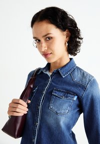 Camicia di denim blu scuro con due tasche frontali, design con bottoni e cuciture a contrasto. Tiene una borsa in pelle bordeaux.