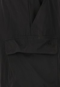 Musta vedenpitävä takki, jossa on läpällinen tasku, sileä pinta ja kulmassa kolmion muotoinen logo. Yksinkertainen, toimiva muotoilu.