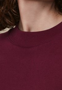 Burgunderfarbener Sweatshirt mit geripptem Rundhalsausschnitt und weicher Stofftextur, mit sichtbarem Nähen entlang des Kragens.