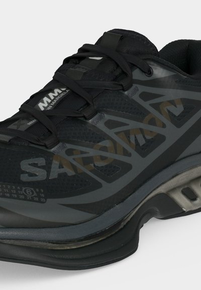 Μαύρο και γκρι αθλητικό παπούτσι με επάνω μέρος από δίχτυ, παχιά μαξιλαρωτή σόλα, μαύρα κορδόνια και λογότυπο "MM6 Maison Margiela" στη γλώσσα και στο πλάι.