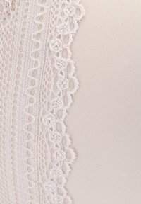 Čipkový lem na mäkkej béžovej tkanine s zložitými kvetinovými vzormi a textúrovaným povrchom, zdôrazňujúci jemné detaily pozdĺž okraja.