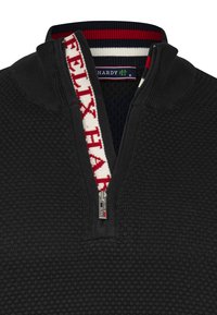 Sudadera negra con cremallera y tejido texturizado, que presenta un cuello alzado y acentos en rojo y blanco en el cuello y la cremallera. Etiqueta de marca visible.