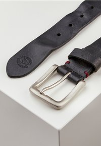 Schwarzer Leder Gürtel mit silberner Metallschnalle, ausgestattet mit einer strukturierten Oberfläche und rotem Stoffdetail. Graviertes Logo auf dem Leder.