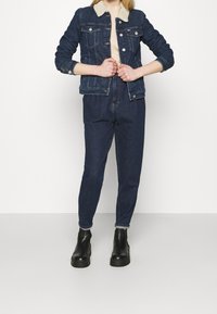 Woman wearing dark denim jacket with fur collar, beige sweater, high-waisted dark jeans, and black platform ankle boots standing against white background.