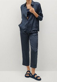 Donna che indossa un completo pigiama a fantasia blu scuro con pantaloni cropped e camicia aperta abbottonata, abbinato a sandali blu navy.