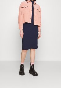 Różowa krótka kurtka z sztruksu z dwoma kieszeniami na piersi, zestawiona z granatową dopasowaną sukienką i czarnymi masywnymi botkami za kostkę z różowymi skarpetkami.