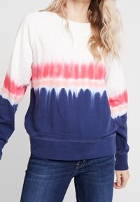 Femme portant un sweat-shirt à manches longues tie-dye en blanc, rouge, rose et bleu marine avec un jean bleu foncé, debout devant un fond uni.